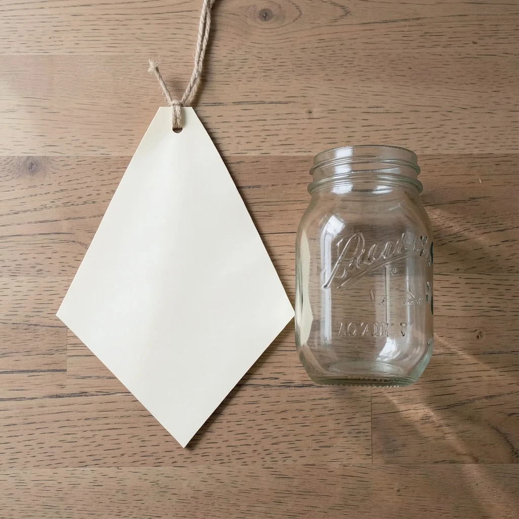 Photograph: A blank cream paper pennant hangs beside an empty clear glass mason jar, with soft window light casting gentle shadows on a neutral background.